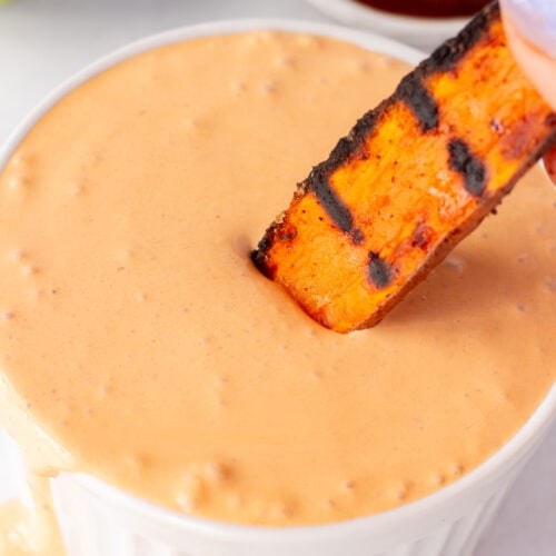 Close up of a grilled sweet potato fry being dipped into an orange sauce in a white bowl.