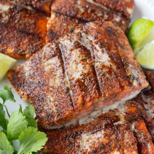 Close up of grilled chili lime salmon filets on a white platter with lime wedges and fresh herbs next to them.