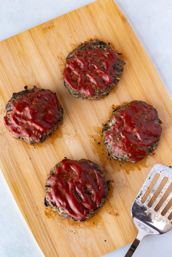 Top down shot of four meatloaf burgers with ketchup on them on a wood cutting board next to a metal spatula.