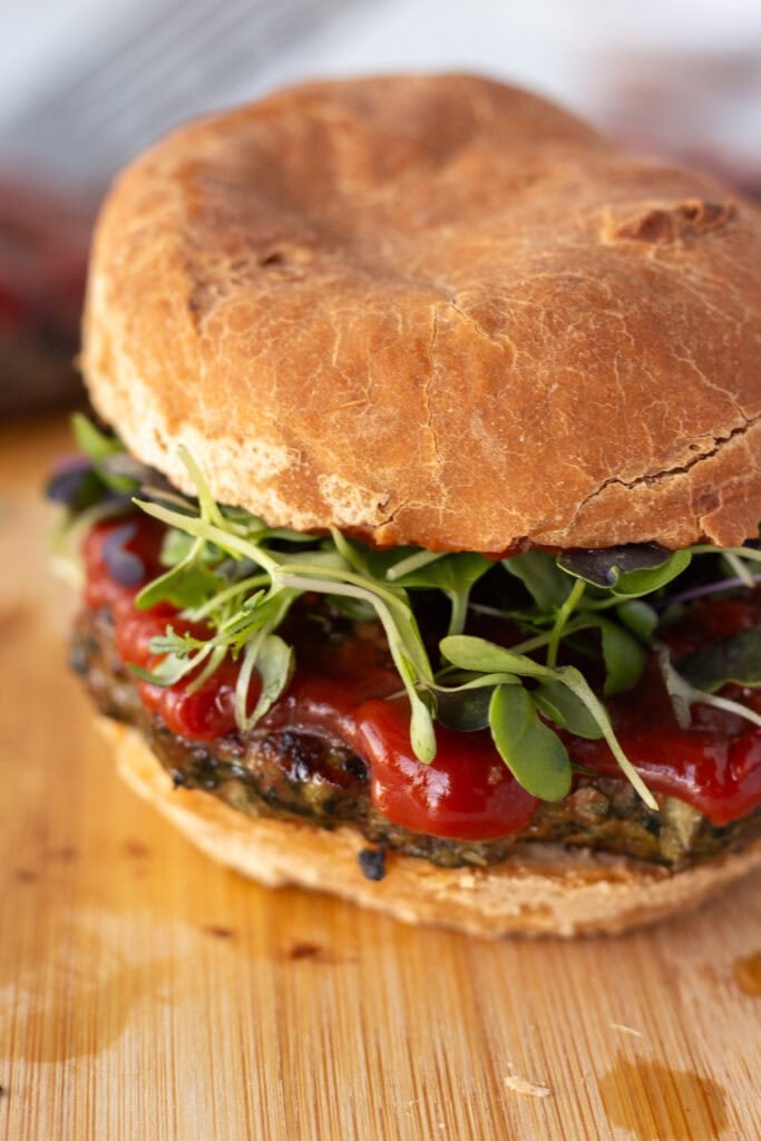 Side close up of a grilled meatloaf burger with ketchup and greens on it , resting on a wood cutting board.