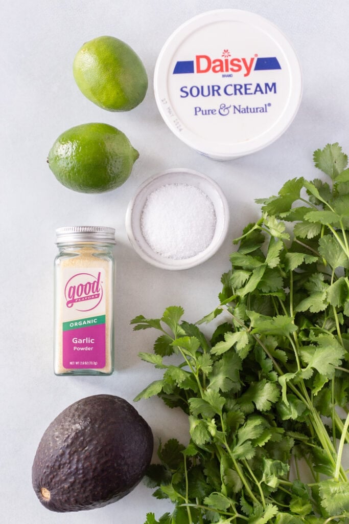 Top down shot of ingredients for cilantro lime sauce, including garlic powder, sour cream, limes, salt, an avocado, and fresh cilantro.