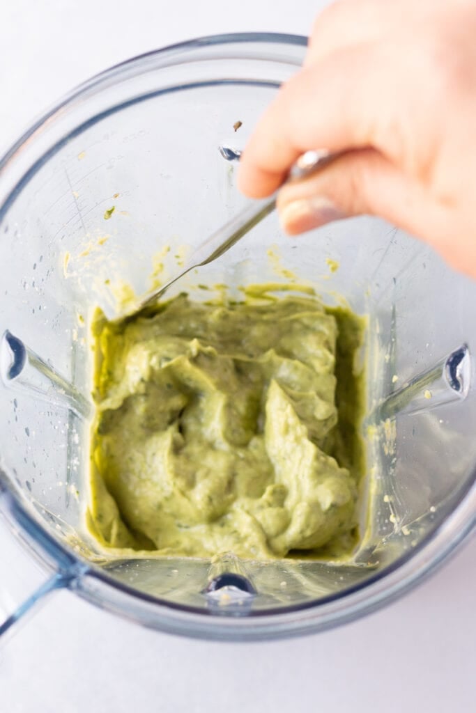 A hand using a butter knife to scrape the sides of a blender with a green avocado cilantro sauce in it.
