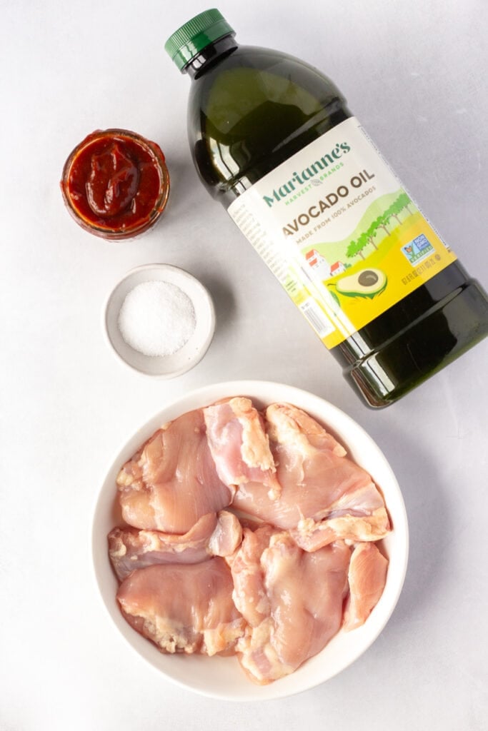 Top down shot of ingredients for bbq grilled chicken thighs, including a bottle of avocado oil, a small bowl with salt, a small jar with bbq sauce, and a white plate with raw chicken thighs on it.