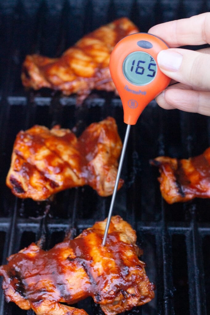 Close up of an orange meat thermometer being stuck in a bbq boneless skinless chicken on a grill. The thermometer reads an internal temp of 165F for the chicken thigh.