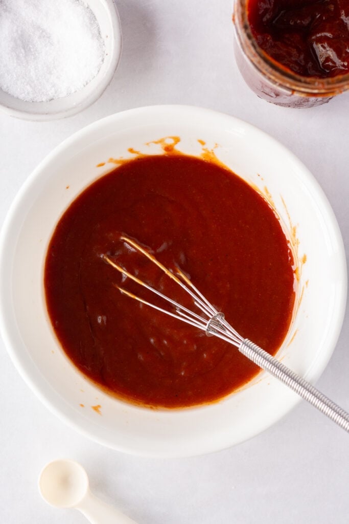Top down shot of a red bbq sauce marinade in a white bowl with a small whisk resting in the bowl.