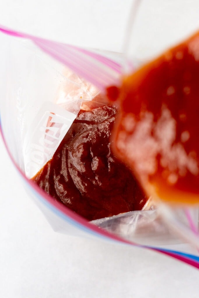 Close up of pouring a red bbq sauce marinade into an open bag that's filled with raw boneless skinless chicken thighs.