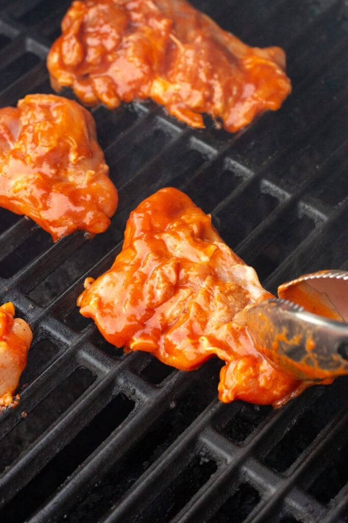A pair of tongs placing raw bbq-marinated boneless skinless chicken thighs on a hot grill.