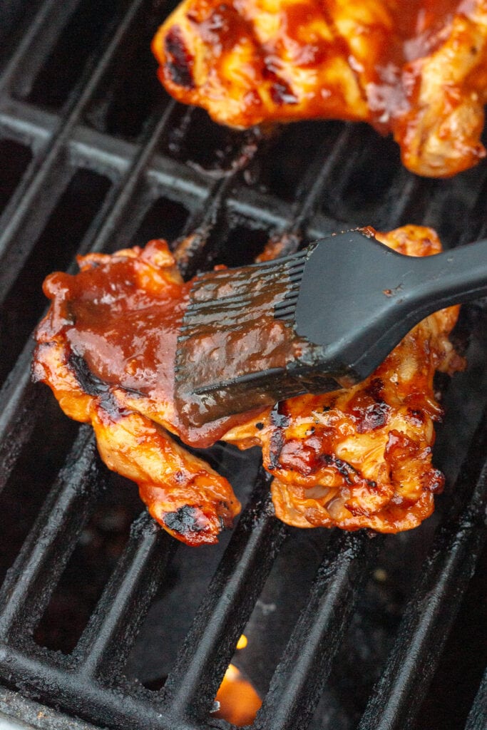 a black basting brush brushing bbq sauce onto a partially cooked boneless skinless chicken thigh on a hot grill.