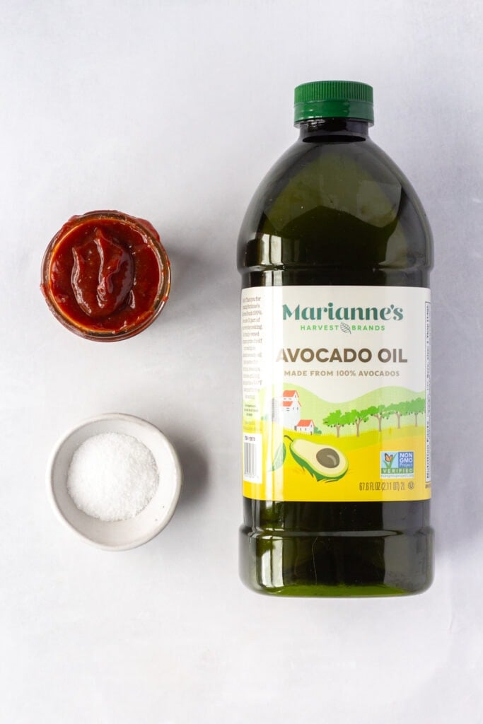 Top down shot of a large bottle of avocado oil, a small bowl of kosher salt, and a small jar of bbq sauce on a light gray background.