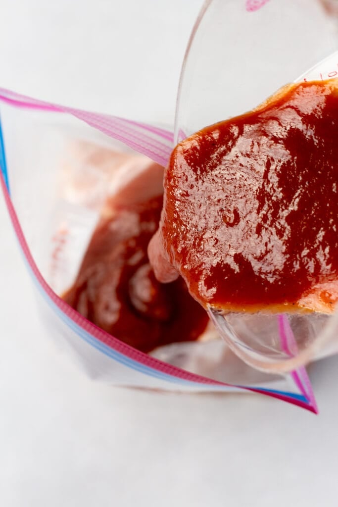 Close up of red bbq marinade being poured into a ziploc bag with raw chicken in it.