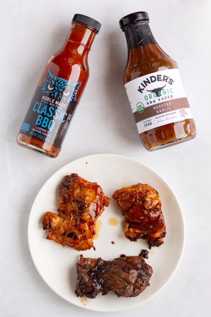 Top down shot of three different pieces of grilled chicken on a white plate next to 2 different bottles of bbq sauce on a light gray background.