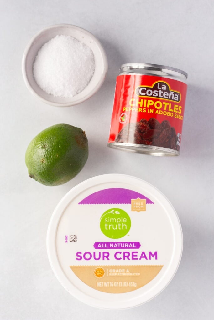 Top down shot of four ingredients for chipotle lime sauce, including sour cream, a lime, a can of chipotle peppers in adobo sauce, and kosher salt.