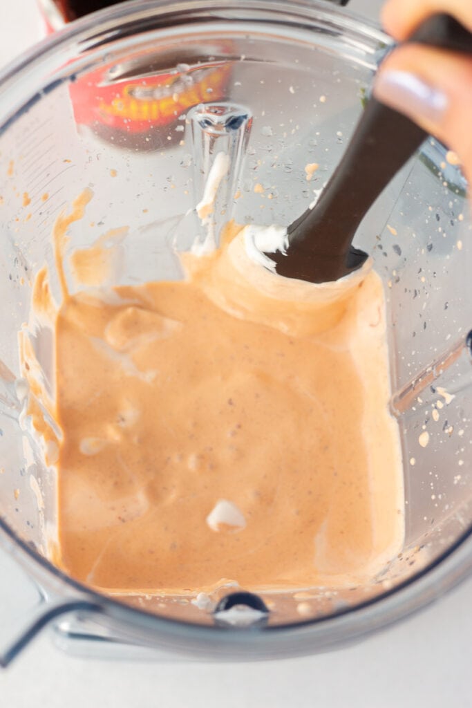 A spatula scraping down the sides of a blender with an orange sauce in it.