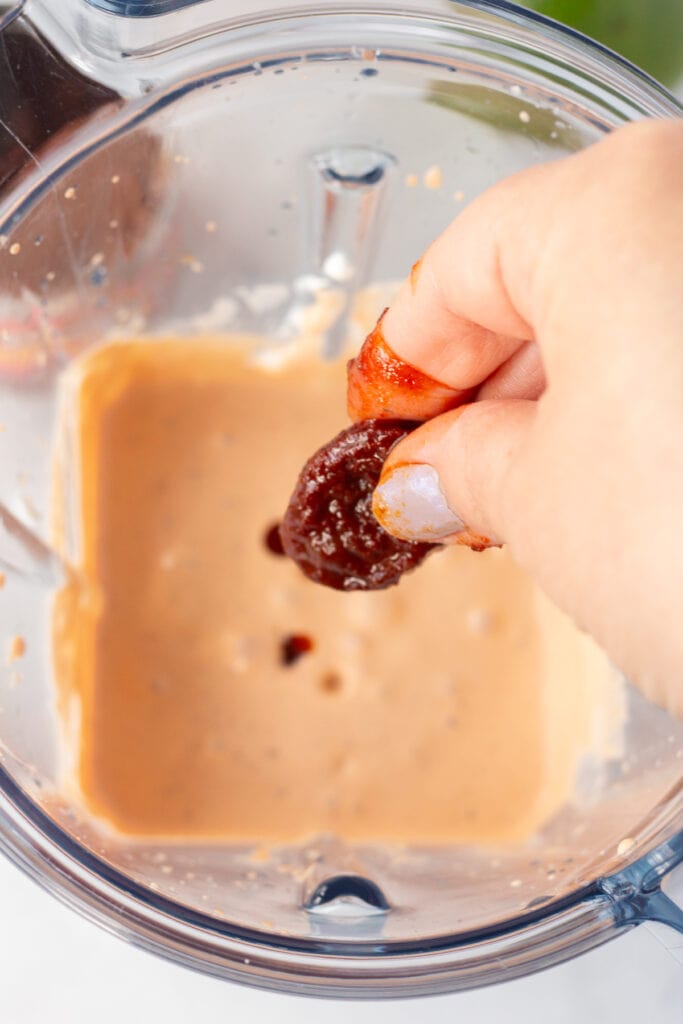 Close up of a hand dropping a chipotle pepper into a blender with an orange chipotle sauce already in it.