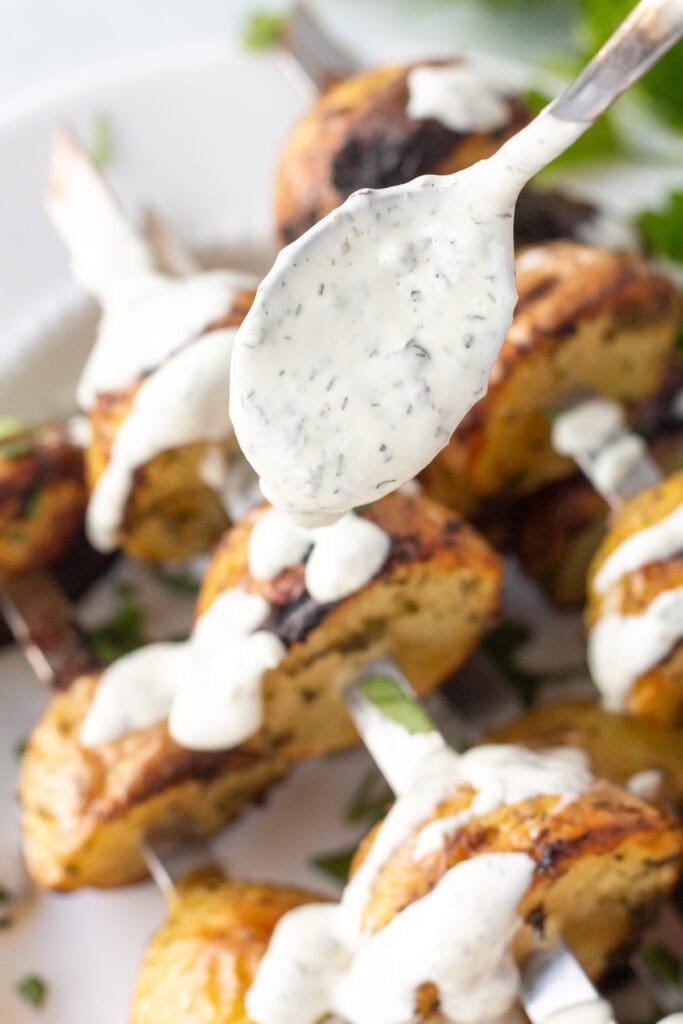 Close up of a ranch dressing being drizzled on grilled potato skewers resting on a white platter.