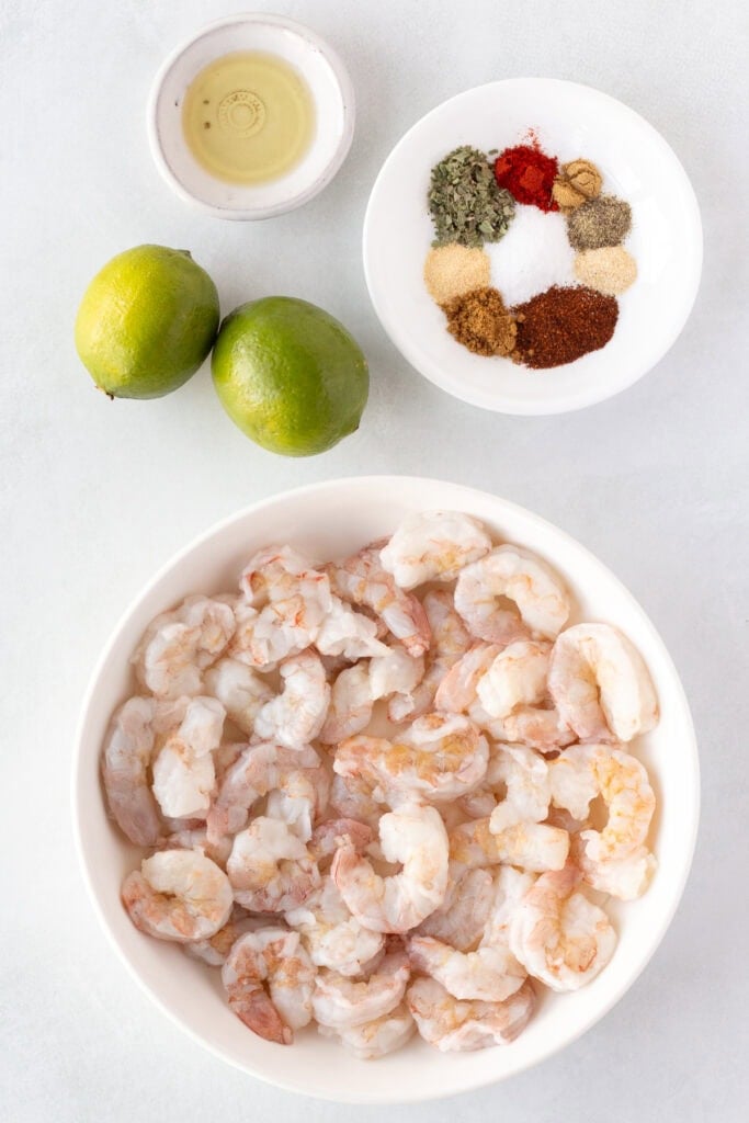 Top down shot of ingredients for chili lime shrimp, including a white bowl with raw shrimp, two limes, a small bowl of oil, and a small plate with several seasonings in it.