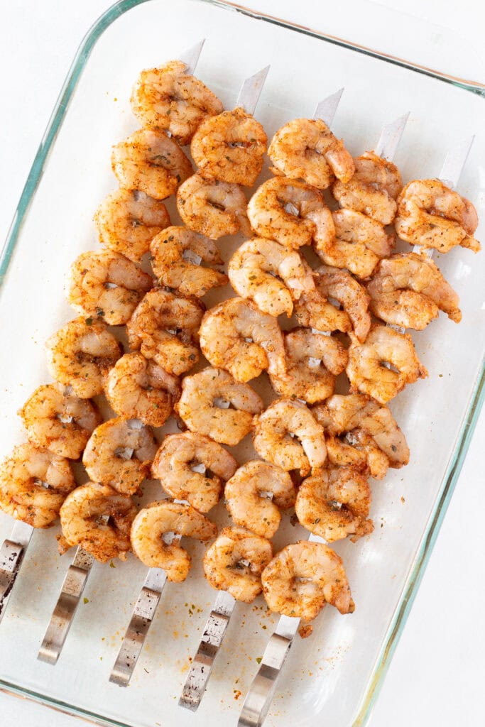 Top down shot of raw shrimp on metal skewers resting in a glass casserole dish.