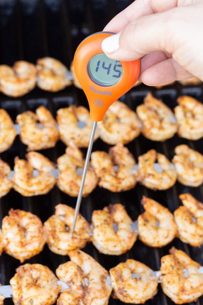 Close up of an orange round temperature probe showing 145F in the digital display as it's stuck in cooked shrimp on metal skewers on a grill.