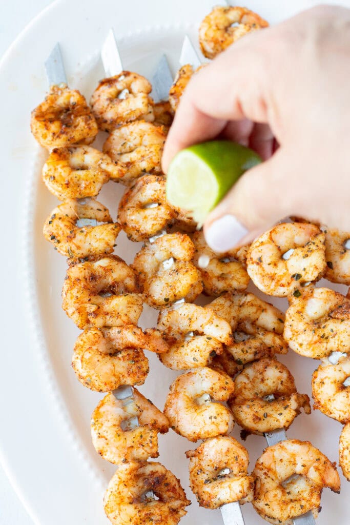 A wedge of lime being squeezed over a white platter with grilled shrimp skewers on it.