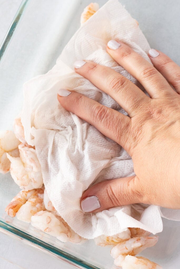 Close up of a hand blotting some raw shrimp in a glass dish with paper towels.