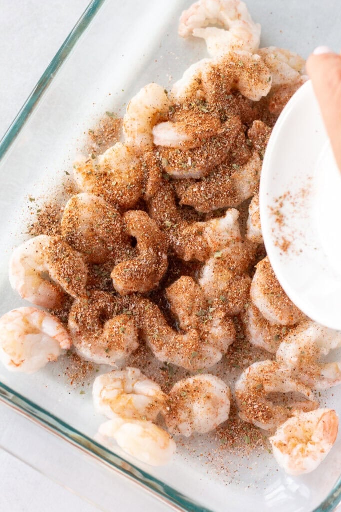 Close up of a chili seasoning being sprinkled onto raw shrimp in a glass dish.