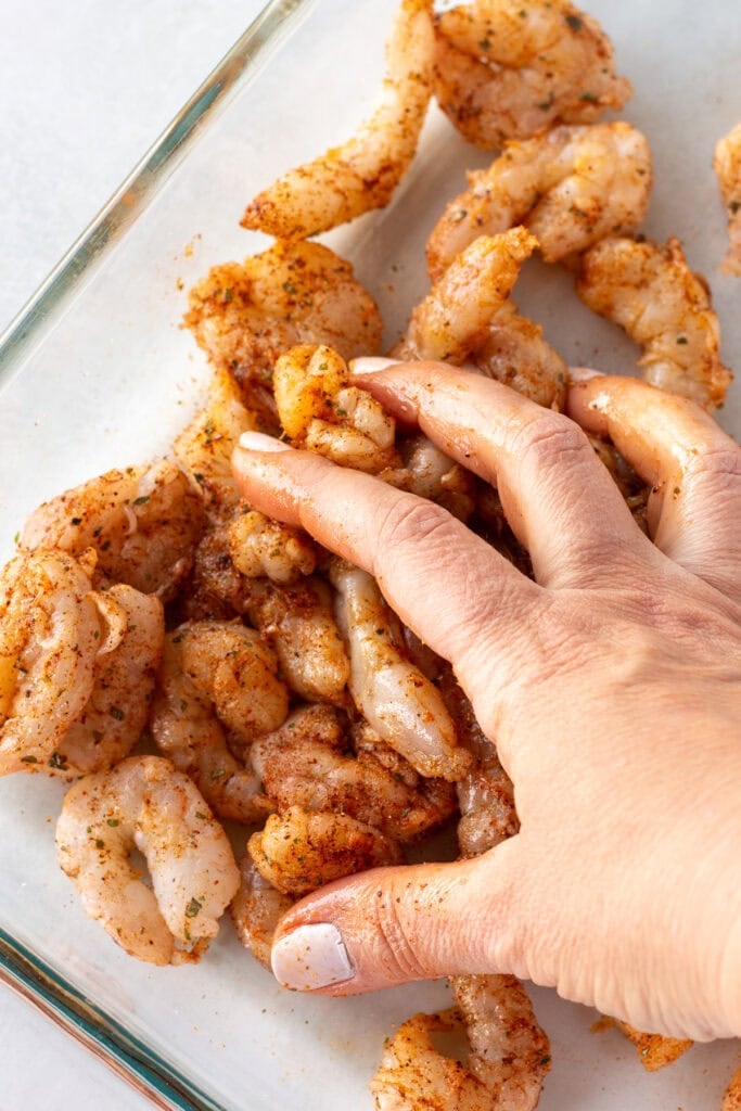 Close up of a hand mixing seasoning and oil onto raw shrimp in a glass dish.