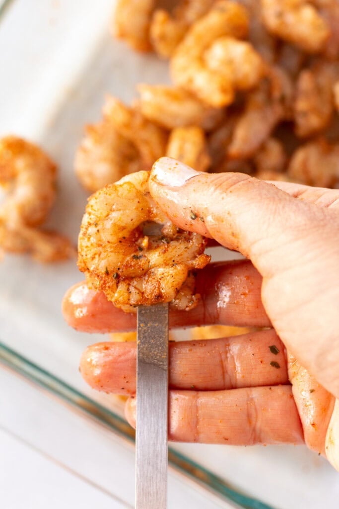 Close up of a hand placing a single raw shrimp on a flat metal skewer.