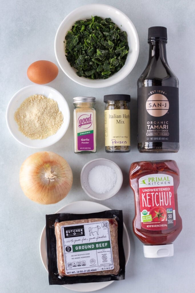 Top down shot of ingredients for grilled meatloaf burgers, includes spices, ketchup, spinach, ground bef, onion, egg, almond flour, and tamari sauce.