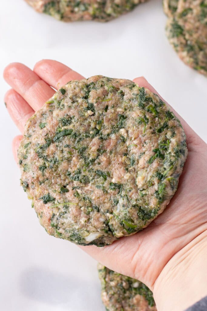 Close up of a hand holding a raw patty of a meatloaf burger with spinach in it.