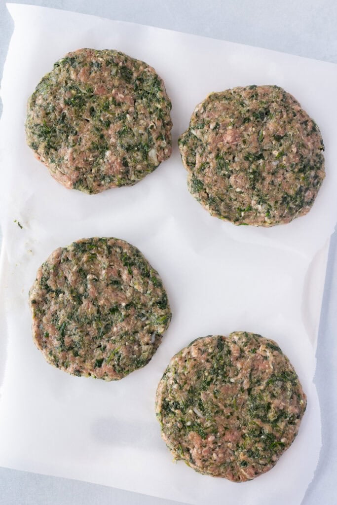 Top down shot of four raw meatloaf burger patties with bits of spinach in them on a white cutting board covered with parchment paper.