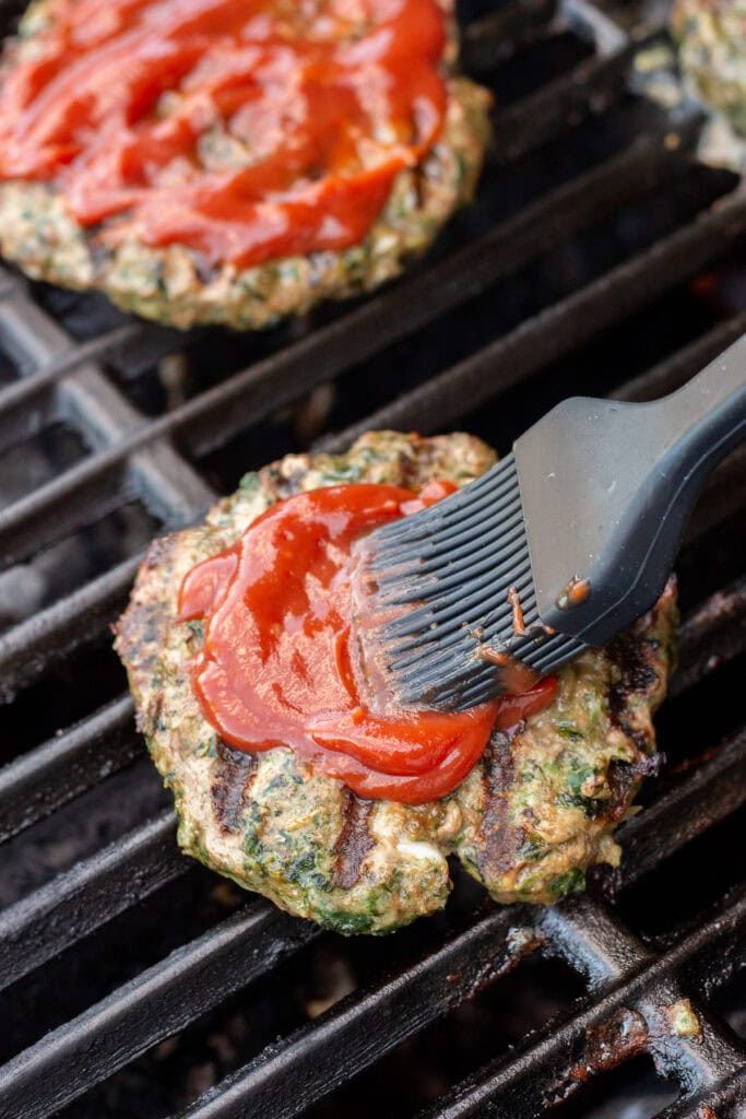 Close up of a basting brush brushing ketchup onto grilled meatloaf burgers on a hot grill.