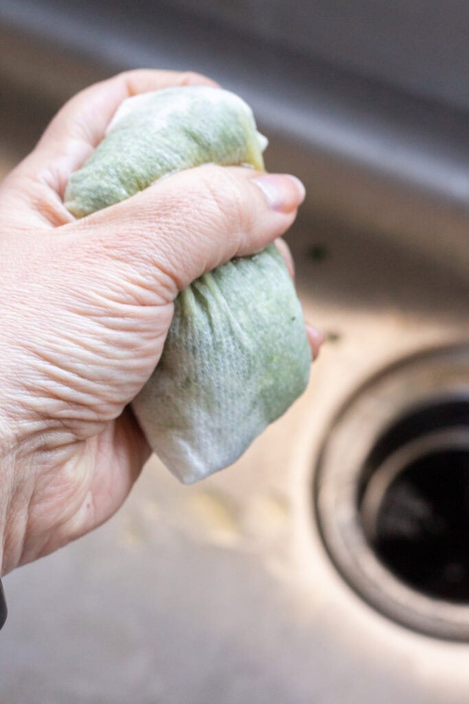 A hand squeezing excess water out of spinach that's wrapped in paper towels over a sink.