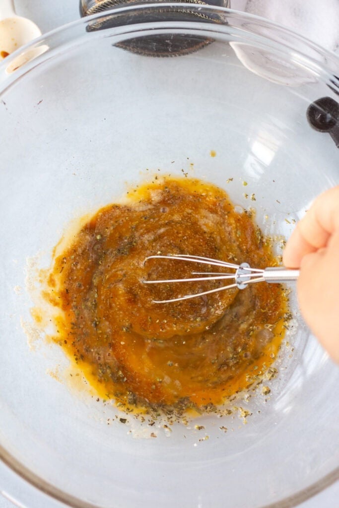 A small whisk stirring together almond flour, raw egg, and spices in a large glass bowl on a light gray surface.