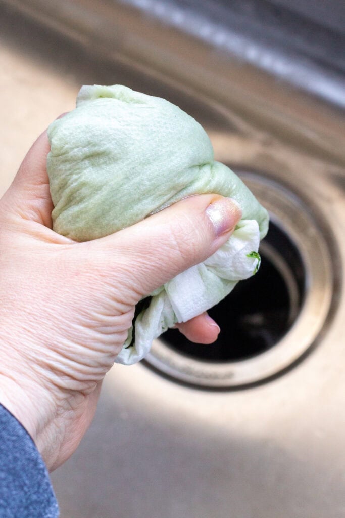 A hand squeezing excess water out of chopped onion and spinach that are wrapped in paper towels. The mixture is held over a sink.
