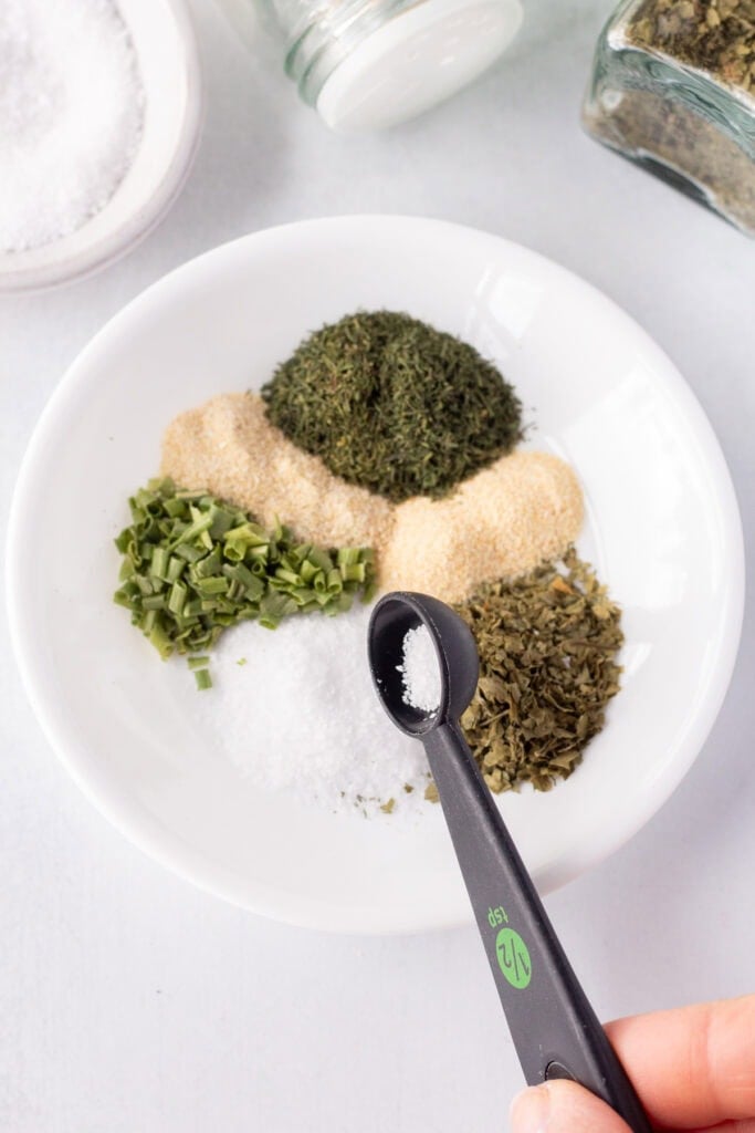 A black measuring spoon pouring kosher salt onto a small plate with other seasonings on it.