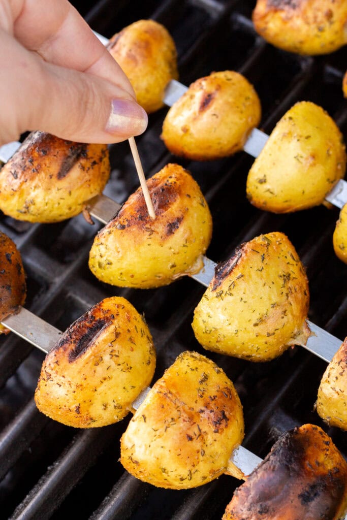 Close up of a toothpick being inserted into halved grilled yellow potatoes on skewers
