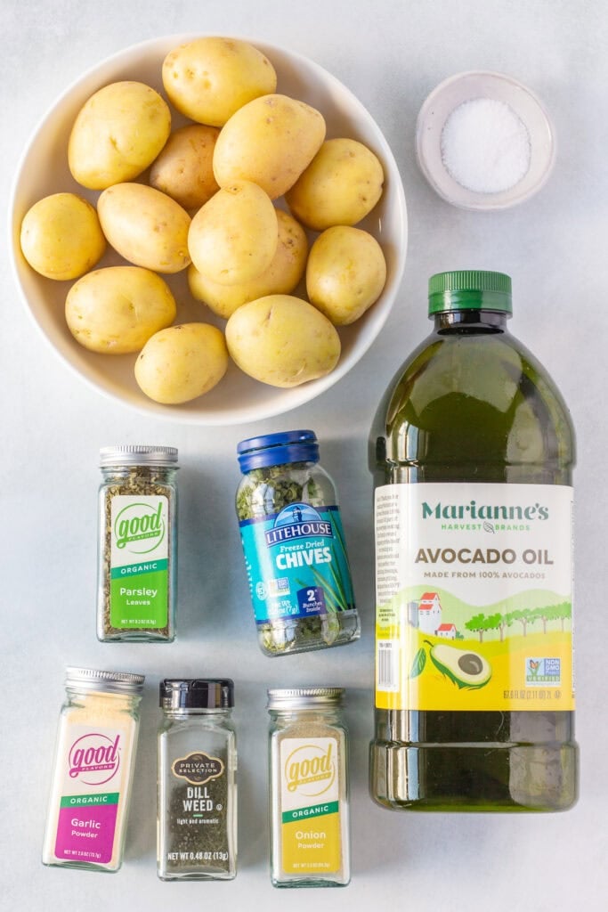 Top down shot of ingredients for grilled ranch potatoes, including a bowl of yellow potatoes, a bottle of oil, and bottles of herbs and spices on a light gray background.