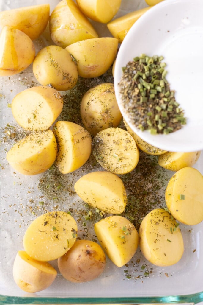 Halved yellow potatoes in a clear flat glass dish with ranch seasoning being poured over them.